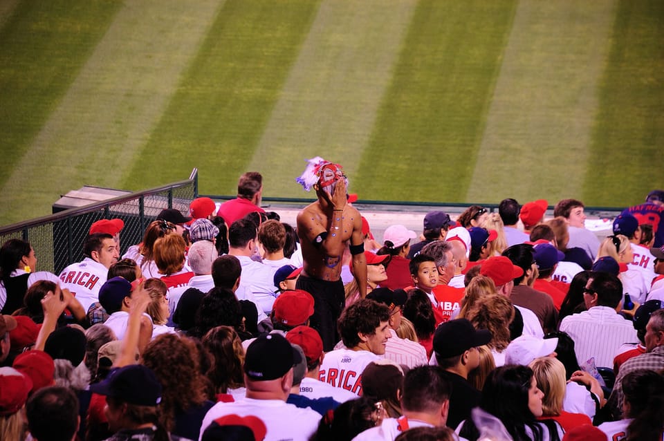 Sports fans in stadium watching the game while another fan, dressed I. Mock face paint and headdress, stands and does a stereotypical Indian yell by patting his hand to his mouth. 
